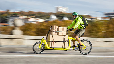 Ciclista su una cargo bike gialla, carica di scatole di cartone fissate davanti, mentre pedala in città indossando casco e abbigliamento sportivo.